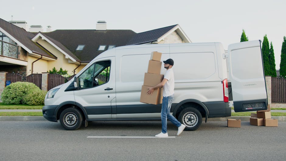 Inside the open rear of a large moving van parked on a paved street, two young women are seated on the edge of the van's loading area, engaging in conversation and smiling. One woman has curly hair, i